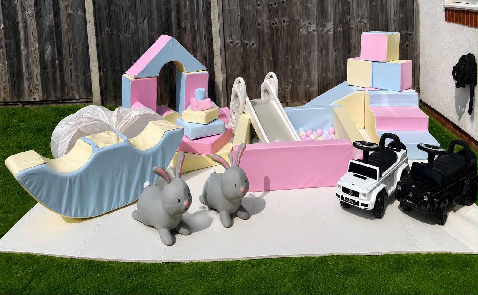 A pastel soft play setup with ball pit, slide and ride-on cars on a garden lawn
